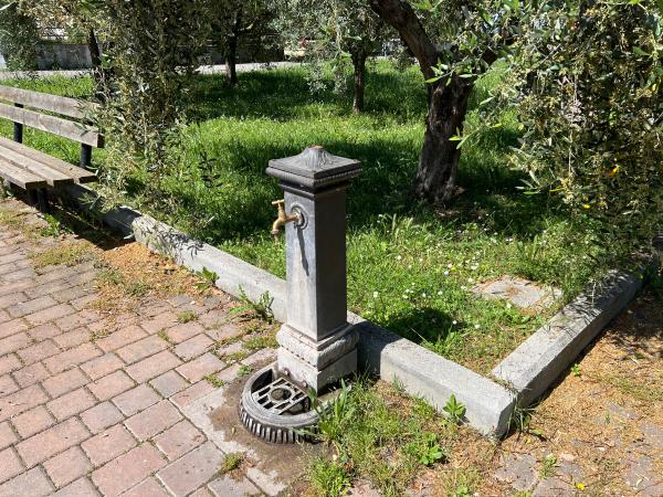 Fontaine métallique avec robinet en or dans le parc de Torricella. Trottoir en briques rouges. Espace vert et banc.